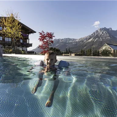 A boy swims in a clear pool with a view of the mountains. In the background, a traditional wooden house and autumn trees can be seen.