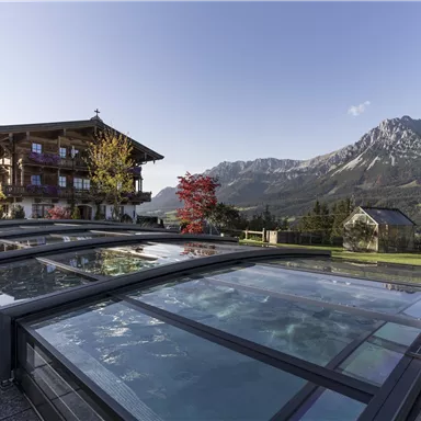 A charming wooden house with a view of majestic mountains. In the foreground, there are modern swimming pools with glass covers.