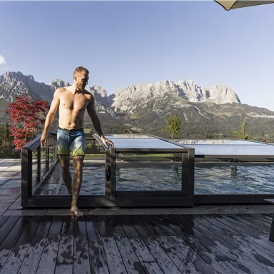 A man stands by a pool with a view of impressive mountains. The landscape is clear and nature appears inviting.