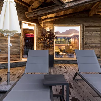 A cozy terrace with loungers and sunshades. In the background are large windows with a view of the mountains.