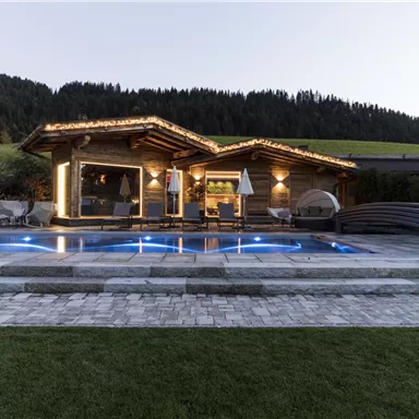 A modern chalet with a illuminated pool. In the background, mountains and green meadows can be seen.