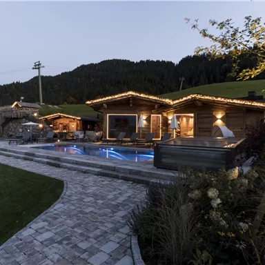 A cozy wooden cabin with warm lights and an outdoor pool. The surroundings are green and mountainous under a clear evening sky.