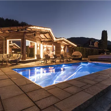 A cozy chalet with a illuminated outdoor area and a swimming pool. The surroundings showcase gentle hills and a tranquil evening atmosphere.
