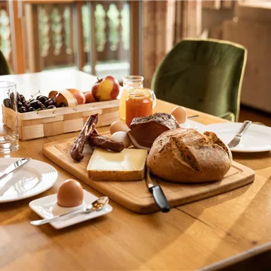 A hearty breakfast on a wooden table with fresh bread, cheese, eggs, and fruit. There are also juices and glasses.