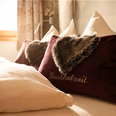 A cozy bed with white pillows and decorative heart-shaped cushions. The warm rays of light create an inviting atmosphere.