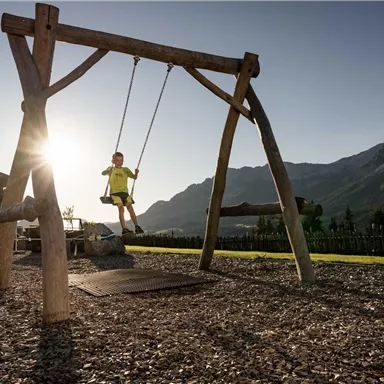 A child swings on a wooden swing outdoors. In the background, mountains and a clear sky are visible.