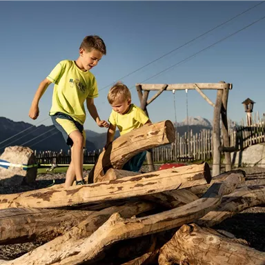 Two children are playing on a pile of wood outdoors. In the background, mountains and playground equipment can be seen.