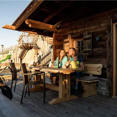 Two children are sitting at a wooden table in front of a rustic mountain hut. They are enjoying drinks in the evening sun.