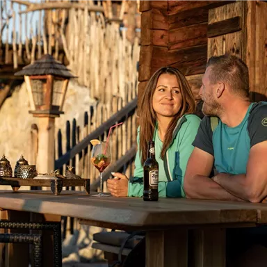 A couple sits relaxed at a table in front of a rustic wooden cabin. They smile at each other and enjoy their drinks in a warm evening atmosphere.