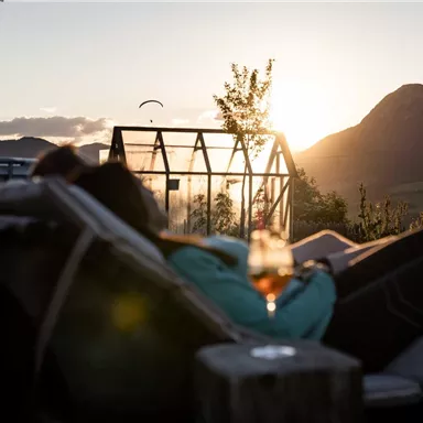 A relaxed person is lying on a lounger and enjoying the sunset. In the background, mountains and a greenhouse can be seen.