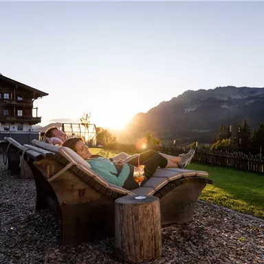 Two people are relaxing in deck chairs as the sun rises. In the background, mountains and a wooden house are visible.