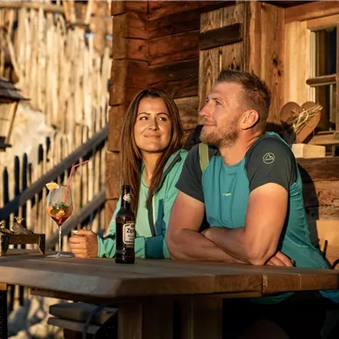 A couple sits at a table in front of a cozy cabin. They enjoy drinks and laugh together in the evening sun.