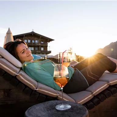 A woman relaxes on a lounger and enjoys a drink. In the background, there is a cozy building and the sun is shining on the horizon.