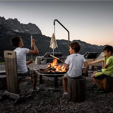 Three children are sitting around a campfire enjoying the evening. In the background, mountains and a clear sky can be seen.