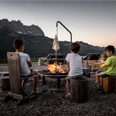 Three children are sitting around a campfire, enjoying the evening. In the background, impressive mountains rise up under a clear sky.