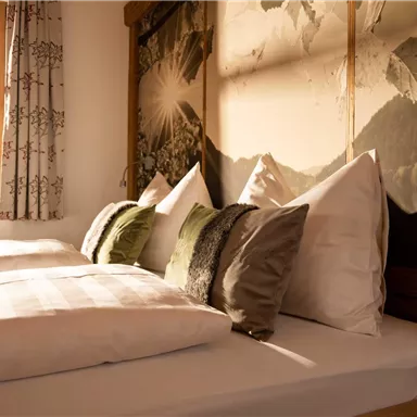 An inviting bedroom with two beds and various pillows. Soft light falls through the window onto the mountain landscape on the wall.