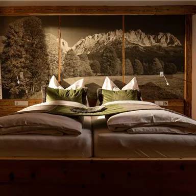 A cozy bedroom with a large double bed and a mountain landscape as wall decoration. The pillows are in various shades of green, creating a relaxing atmosphere.