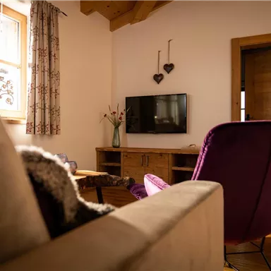 A cozy living room with a beige sofa and a purple chair. The walls are light, and there is a window with curtains letting natural light in.