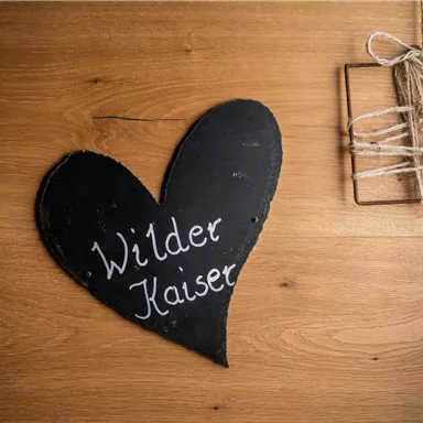 A black heart-shaped sign with the inscription "Wilder Kaiser" hangs on a wooden wall. Next to it, a small decorative wreath is attached.