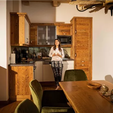 A modern kitchen made of wood with a dining table and cozy chairs. A woman is standing smiling in the kitchen.
