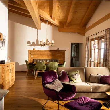 A cozy living space with wooden ceilings and large windows. The room is modernly furnished with a sofa and a dining table.