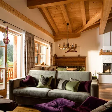 A cozy living room with high wooden beams and a modern fireplace. The sofa is adorned with decorative pillows and large windows let in plenty of light.