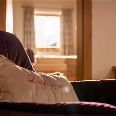 An elegant armchair with a decorative cushion in the foreground. In the background, a bright window with curtains can be seen.