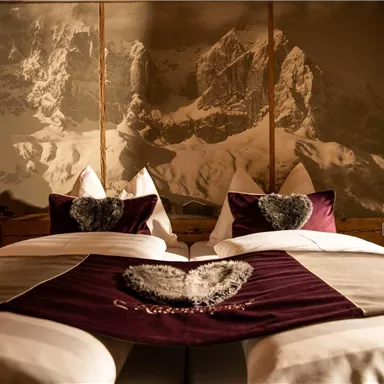 A cozy bedroom with two beds and heart pillows. In the background, there is an impressive mountain landscape.