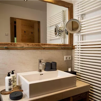 A modern bathroom with a rectangular sink and a wooden frame mirror. On the wall, there are radiators and personal care products placed on the sink.