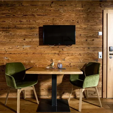 A cozy dining area with a wooden table and two green chairs. On the wall, there is a television and a wooden door.