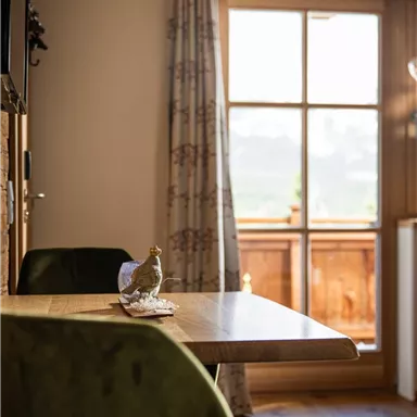 A cozy interior with a wooden table and green chairs. Sunlight is shining through the window, and the view shows a rural landscape.