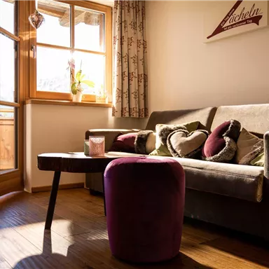 A cozy living room with a sofa and cushions in gentle colors. There are large windows that let in plenty of daylight.