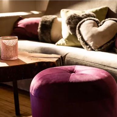 A cozy living room with a gray sofa and colorful cushions. On a table, there are candles and a few round, purple poufs.