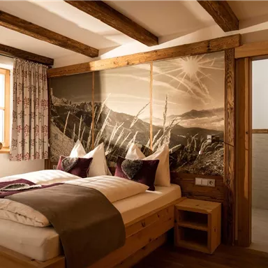 A cozy bedroom with a wooden double bed and a beautiful view of the mountains in the background. The room has large windows and rustic decoration.