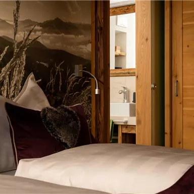 An elegant bedroom with a cozy blanket and pillows in soft colors. In the background, a wooden shelf and an adjoining bathroom are visible.