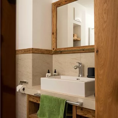 A modern bathroom with a white sink and wooden shelves. Green towels and toiletries are placed on the shelf.