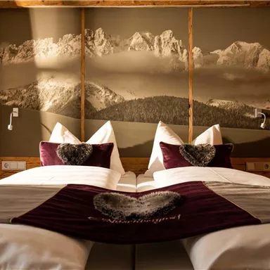 A cozy bedroom with two beds, adorned with white pillows and a red blanket. In the background, there is a large picture of mountains.