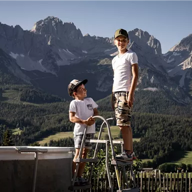 Two boys are standing on a ladder next to a pool, with an impressive mountain landscape in the background. The sun is shining and the sky is clear.