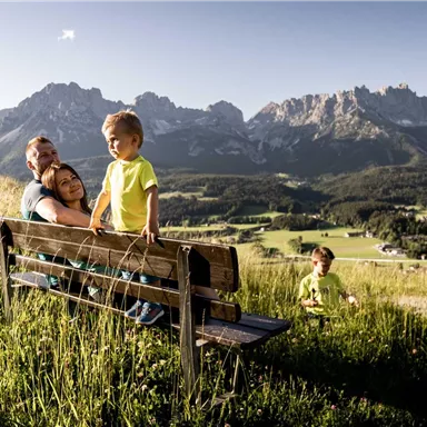 A family is sitting on a bench in nature. In the background, mountains and a green landscape can be seen.