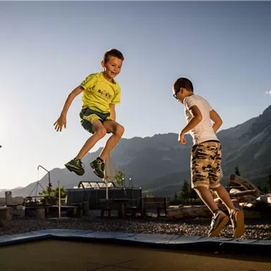 Two children are happily jumping on a trampoline. In the background, mountains and a clear sky are visible.