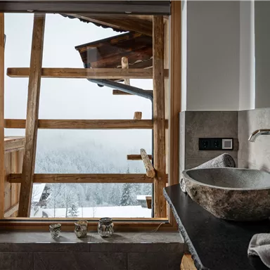 A modern bathroom with a stone washbasin and wooden framed windows. The view shows a snowy landscape with fog.