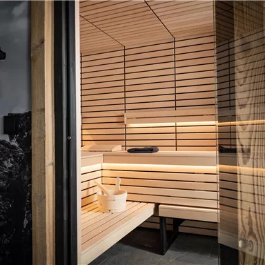 A modern sauna made of light wood with gentle lighting. In the foreground is a bucket for water and a basin that radiates coziness.