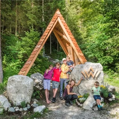 A group of five people stands in front of a wooden entrance with a pointed roof. Around them is a forest with many trees and green vegetation.