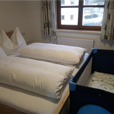 Ein gemütliches Schlafzimmer mit zwei Betten und großen Kissen. In der Ecke steht ein Babybett.