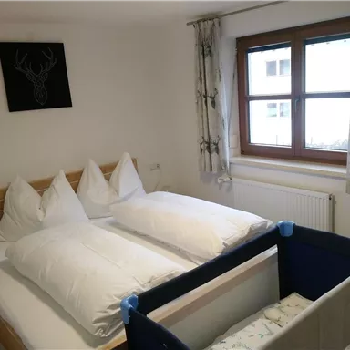 Ein gemütliches Schlafzimmer mit einem Doppelbett und einem Kinderbett. Ein Fenster mit Blick nach draußen und helle Vorhänge sorgen für freundliches Licht.