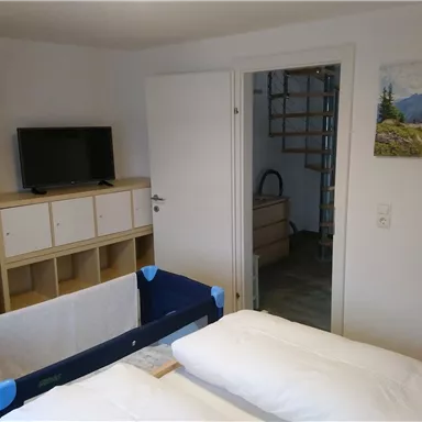 Ein einfaches Schlafzimmer mit einem Fernseher, einem Kleiderschrank und einem Kinderbett. An der Wand hängt ein Bild mit einer Berglandschaft.