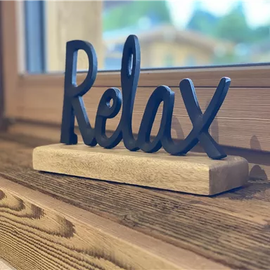 A decorative sign with the word "Relax" made of black material is placed on a wooden table. In the background, a window is visible, allowing a natural glow of light.