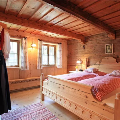 Ein gemütliches Schlafzimmer mit Holzwänden und einem Doppelbett. An der Wand hängt ein traditionelles Kleid und es gibt große Fenster mit Vorhängen.