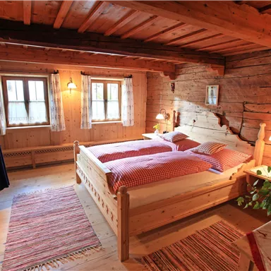 Ein gemütliches Schlafzimmer mit einem Holzbett und roten Bettwäsche. Die Wände sind aus Holz, und es gibt Fenster, die viel Licht hereинlassen.