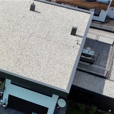 Ein modernes Flachdach mit Kiesbelag und mehreren Lüftungsöffnungen. Im Hintergrund sind weitere Gebäude und Terrassen sichtbar.